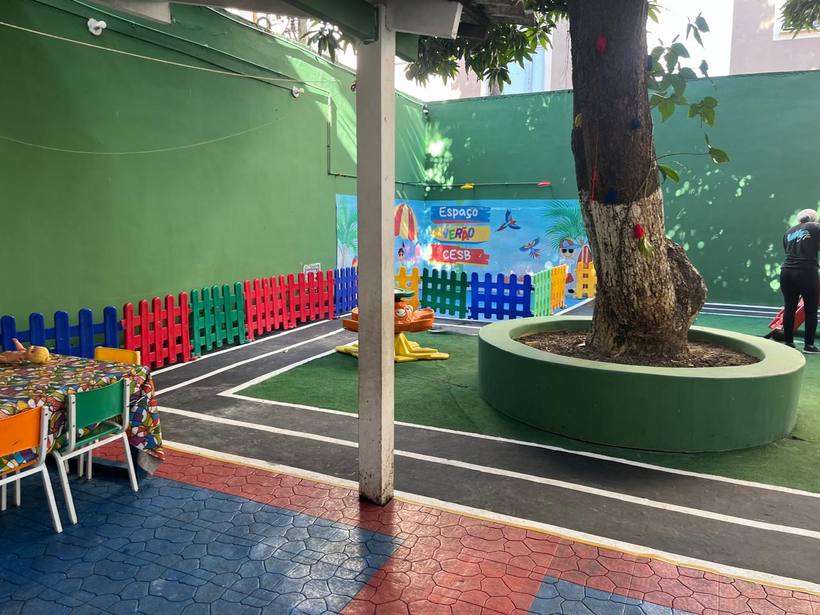 Área de recreação ao ar livre, com mesinhas, brinquedos e sombra de árvore