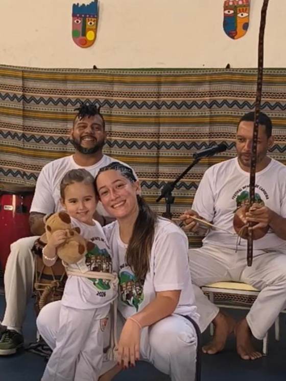 Aluna da Creche Escola Santa Beatriz com a professora de capoeira, ao lado dos músicos que tocam na roda