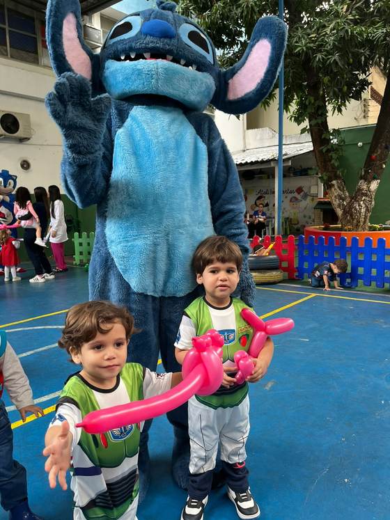 Alunos fantasiados brincando com balões ao lado de um personagem no pátio da escola, no Dia das Crianças