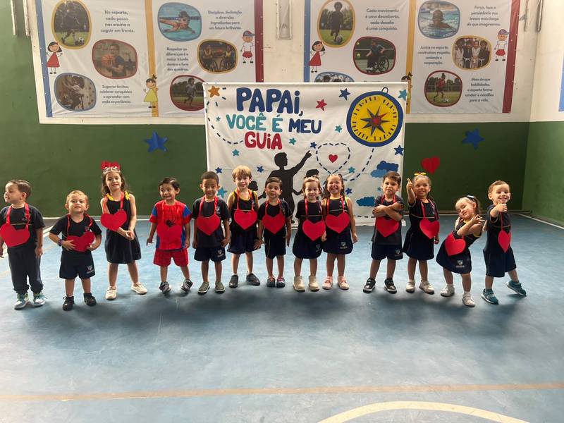 Turma da Creche Escola Santa Beatriz enfileirada com corações de papel em frente ao painel Papai, você é meu guia
