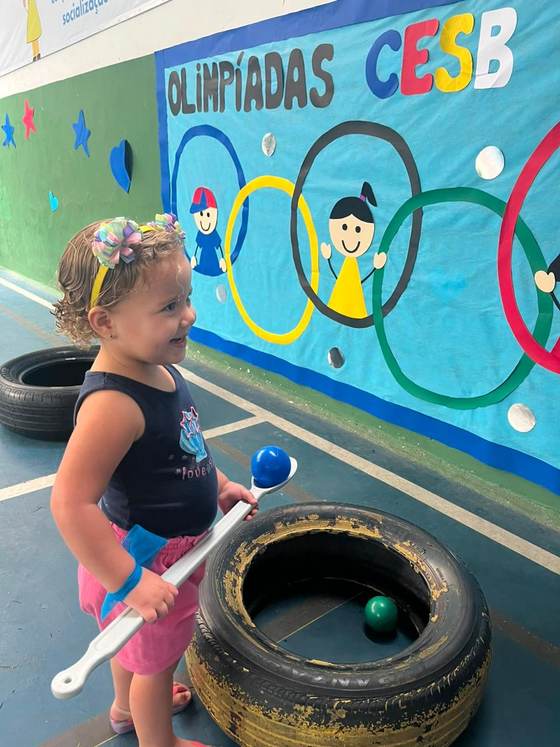 Aluna da Creche Escola Santa Beatriz na prova da bolinha na colher, em frente ao painel das Olimpíadas CESB