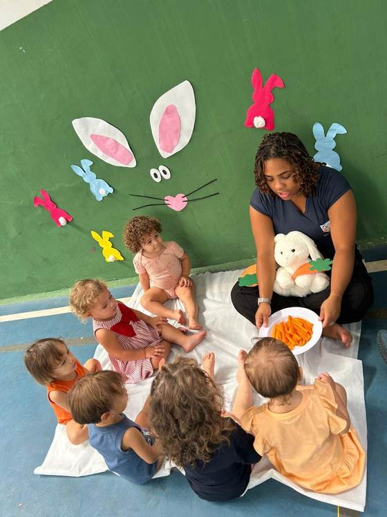 Turma do Berçário da Creche Escola Santa Beatriz em roda de história de Páscoa com a professora, o coelho de pelúcia e o painel de orelhas na parede