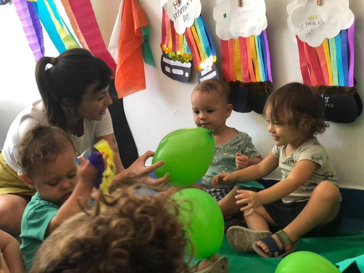 Turma do Berçário brincando com balões verdes em atividade do Saint Patrick's Day, com os arco-íris de papel feitos pelas crianças ao fundo